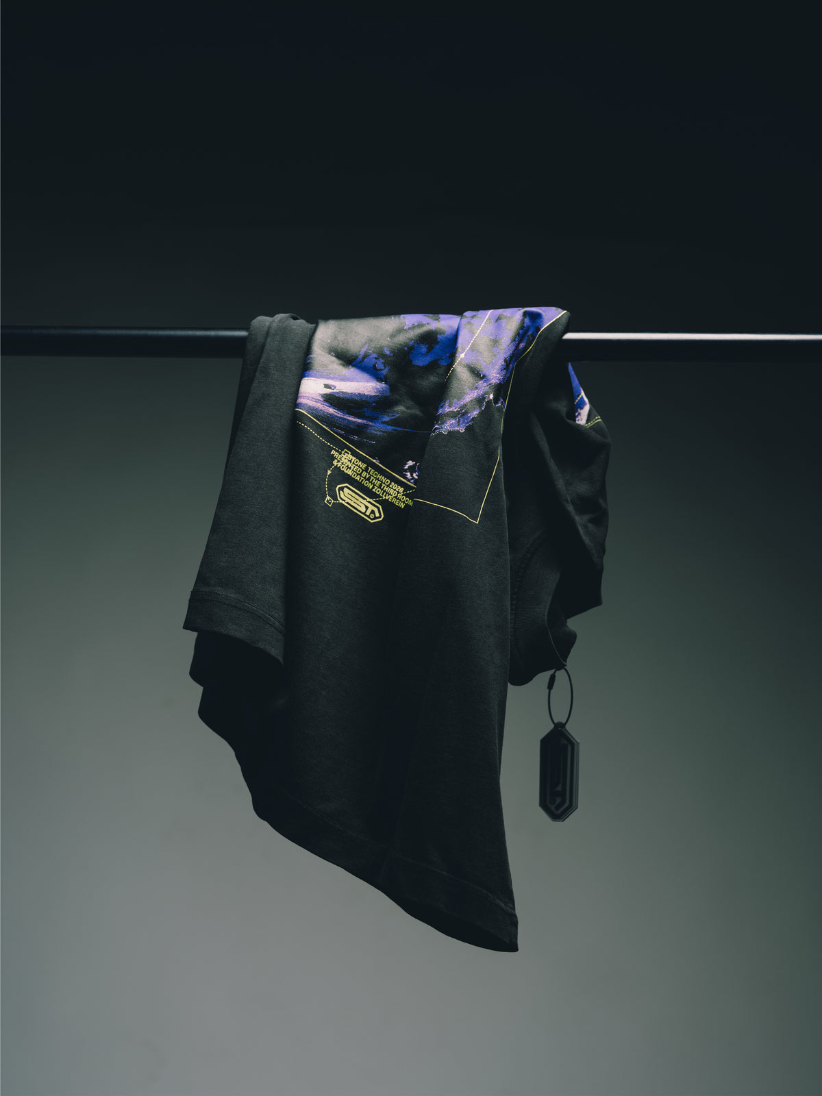 Schwarzes T-Shirt mit blau-violettem Grafikprint und gelben Details, locker über eine Kleiderstange gehängt vor dunklem Hintergrund.