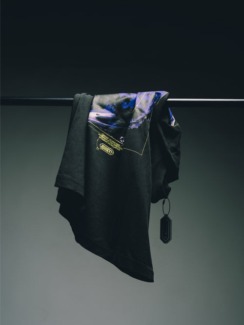 Schwarzes T-Shirt mit blau-violettem Grafikprint und gelben Details, locker über eine Kleiderstange gehängt vor dunklem Hintergrund.