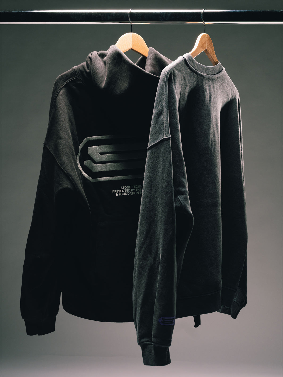 Schwarzer Hoodie und dunkelgraues Sweatshirt auf Holzbügeln vor grauem Hintergrund, mit dezenten Logo- und Printdetails.