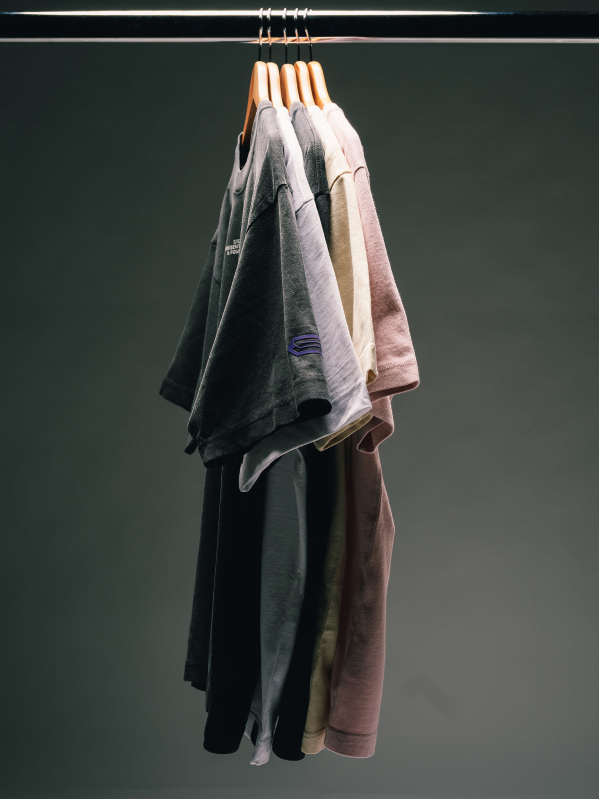 Mehrere T-Shirts in Schwarz, Grau, Beige und Rosa dicht nebeneinander auf Holzbügeln an einer schwarzen Kleiderstange.